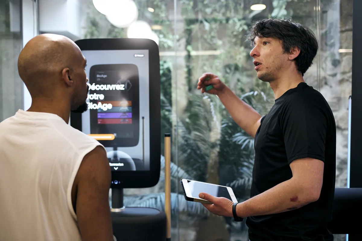 Un coach en t-shirt noir commente les résultats d'un bilan sur une tablette devant une borne tactile affichant « Découvrez votre BioAge », face à un adhérent, site Paris Marais