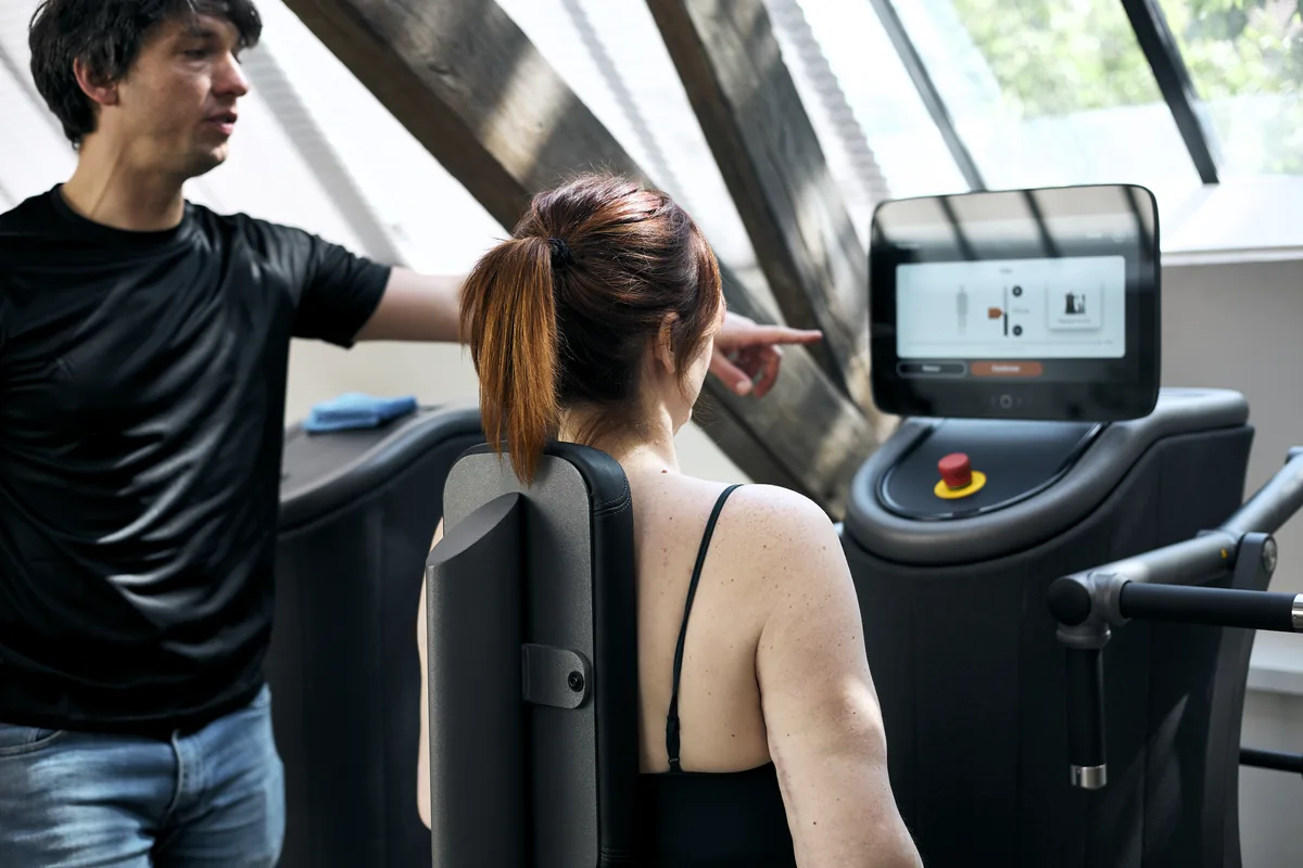Un coach de la Maison du Peps montre du doigt à une adhérente assise le réglage affiché sur l'écran d'une machine eGym, salle du Marais