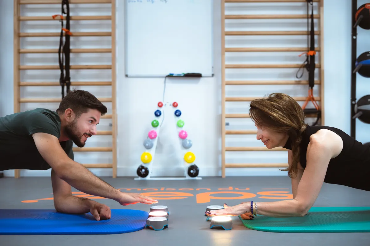 Deux adhérents en gainage face à face touchent des plots lumineux sur leurs tapis