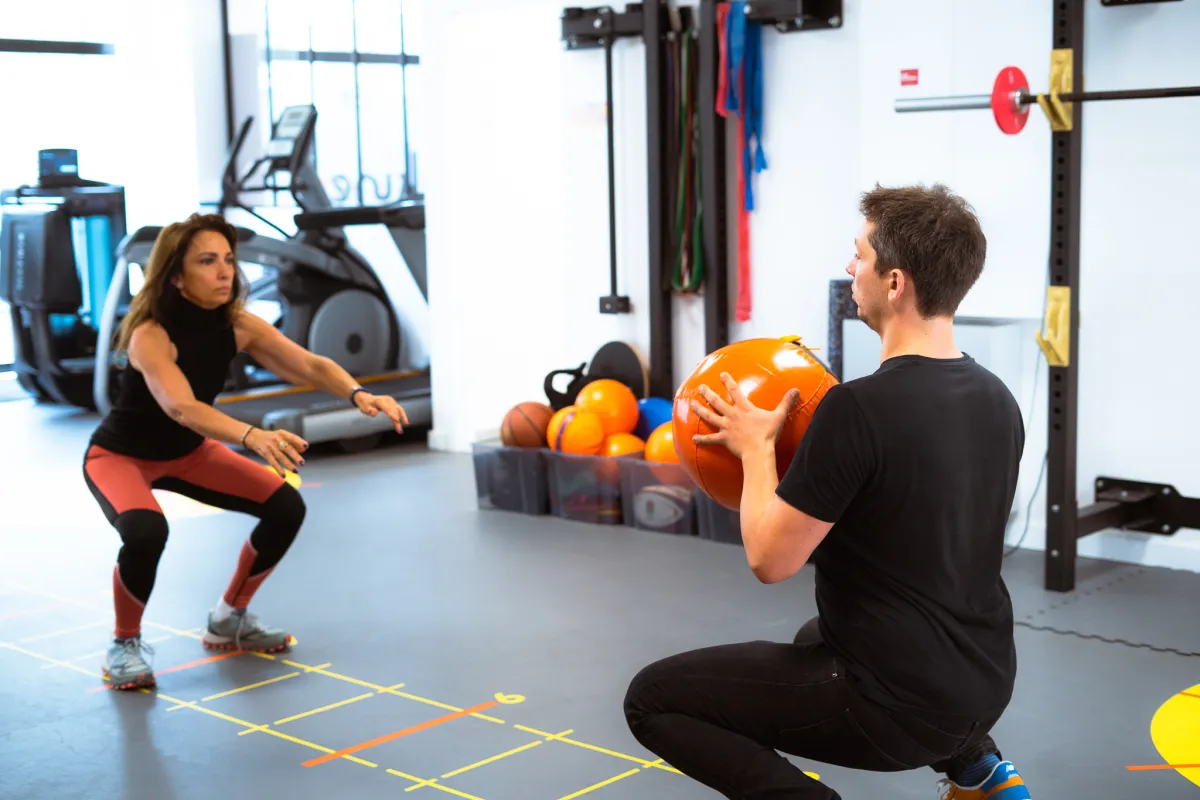 Deux adhérents enchaînent des squats, l'un avec un médecine-ball orange, sur l'échelle d'agilité