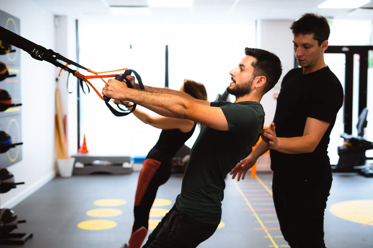 A member performs a TRX suspension row while a trainer supervises his position